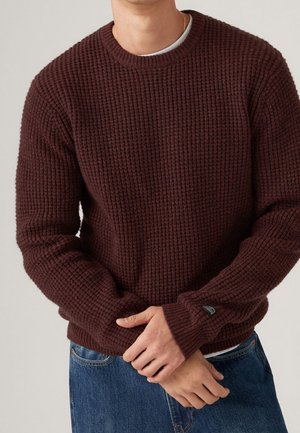 Mand iført brun vaffelstrikket sweater over hvid skjorte, med blå jeans, hænderne foldet foran, neutralt baggrund.
