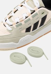 Zapatilla con una parte superior de ante y textil en tonos beige y verde, rayas negras, cordones blancos y cordones verde claro a su lado.