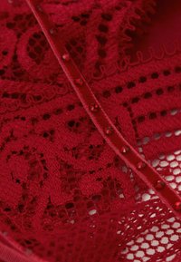 Červená krajka s nádhernými vzory, otevřenými úseky tkaniny a saténovou stuhou zdobenou červenými drahokamy.