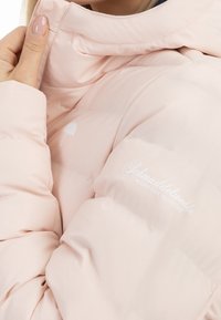 Detailní záběr na osobu, která upravuje límec světle růžového péřového kabátu s bílým deštníkem a nápisy na hrudi a rukávu.