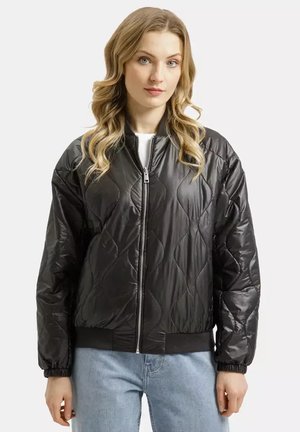 Blondynka ubrana w czarną pikowaną kurtkę na zamek i jasnoniebieskie dżinsy, stojąca na tle gładkiego jasnego tła.