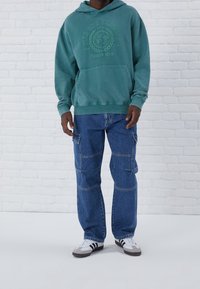 Felpa verde con cappuccio e logo, abbinata a pantaloni cargo blu con molte tasche e una vestibilità comoda, completata da sneaker nere e bianche.