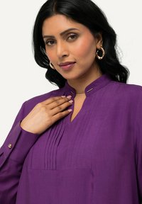 Paarse blouse met een mandarinakraag, voorzien van plooien aan de voorkant en knoopmanchetten. Soepele stof met een lichte glans.