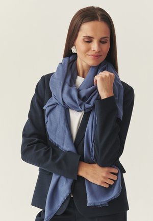Kobieta w granatowej marynarce i białej bluzce, z dużym miękkim niebieskim szalem owiniętym wokół szyi oraz srebrnymi teksturowanymi kolczykami.