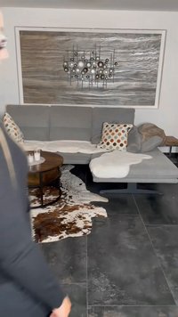 Sofá seccional cinza com almofadas circulares com pontos laranja, castanho e creme, colocado sobre um tapete de pele de vaca em cima de um chão de azulejos escuros.