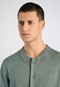 Ljusgrön polo med en texturerad stickning, som har en krage, två knappar och en liten logotyp på vänster sida. Slät tyg.