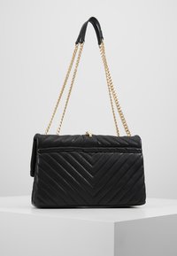 Černá prošívaná kožená kabelka s chevron vzorem, zlatý řetízek s černou koženou podložkou na rameno, položená na bílé povrchu.