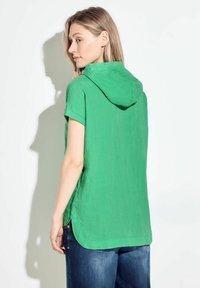 Maglietta verde a maniche corte con cappuccio in tessuto testurizzato, caratterizzata da un orlo curvo e spacchi laterali, abbinata a jeans in denim blu.