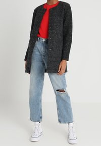 Cardigan lungo grigio carbone con scollatura arrotondata, bottoni a pressione e finitura testurizzata, abbinato a una t-shirt rossa e jeans strappati blu chiaro.