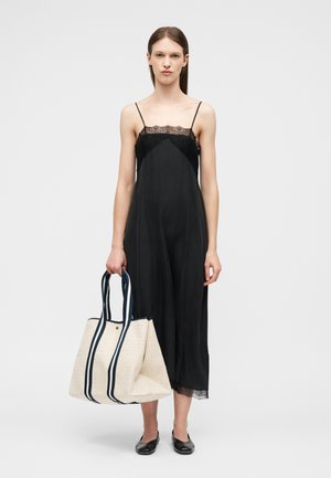 Γυναίκα που φοράει μαύρο φόρεμα με λεπτομέρειες δαντέλας και κρατά μια μεγάλη μπεζ τσάντα tote με λουράκια σε σκούρο μπλε ρίγες.