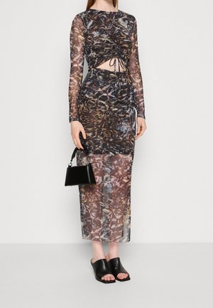 Woman in a long, sheer, patterned dress with ruched front, holding a small black handbag, wearing black open-toe mules.