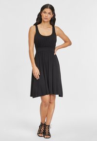 Schwarzes ärmelloses Kleid mit einem taillierten Oberteil und einem fließenden Rock. Hergestellt aus weichem Stoff, verfügt es über eine geraffte Taille und eine glatte Oberfläche.