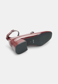 Red patent leather heel with a square shape and ankle strap. Cushioned insole and textured rubber outsole. Features a branded logo on the sole.