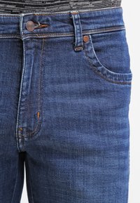 Mörkblå denimjeans med en klassisk femficksdesign, kontrasterande orange sömmar och metalldetaljer på knappen vid midjan.