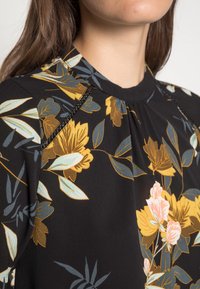 Blouse noire à fleurs avec des fleurs jaunes et bleu clair, encolure plissée et détails de couture contrastante sur les épaules. Texture de tissu douce.