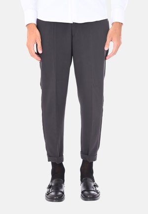 Pantaloni neri con una vestibilità sartoriale, texture subtile, orli arrotolati e linee pulite, abbinati a scarpe slip-on nere e calzini scuri.