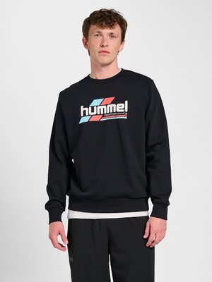 Young man with curly hair wearing a black sweatshirt with a red and blue "hummel performance" logo, standing against a plain white background.