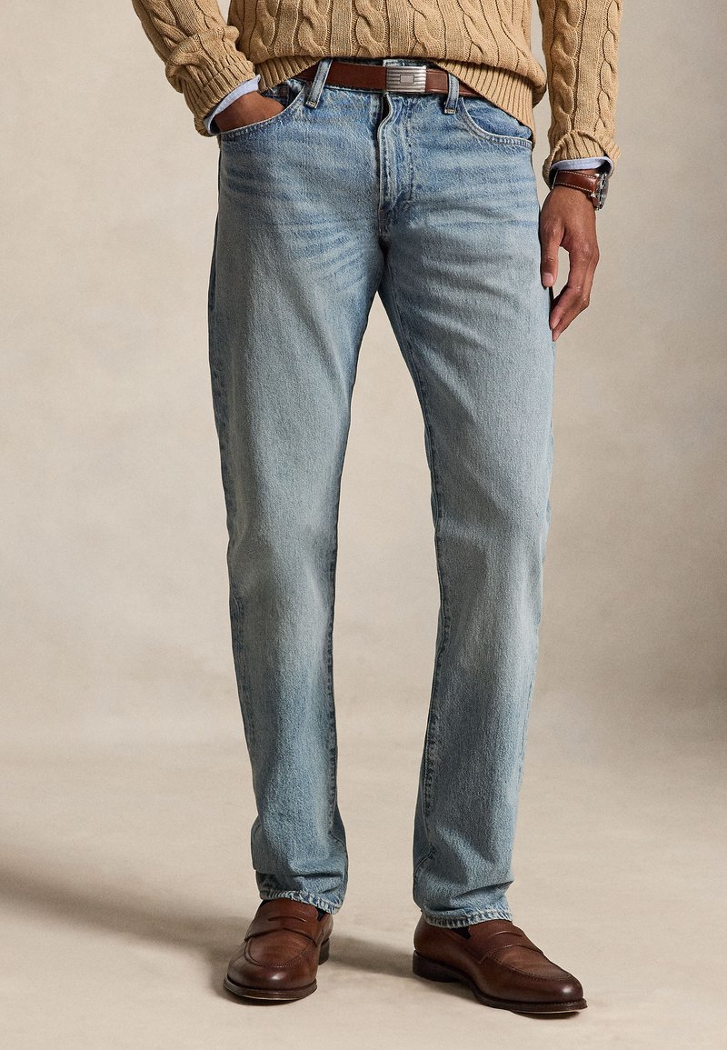 Mand iført lyseblå jeans, brunt læderbælte, brune lædersko, lysebrun kabelstrikket sweater og et armbåndsur, står med afslappede hænder.