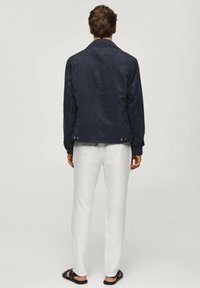 Muž v tmavě modré bundě, bílých kalhotách a černých sandálech, stojící zády k hladkému světlému pozadí.