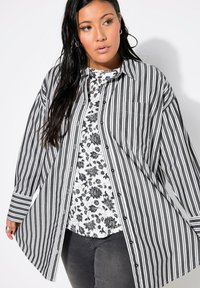 Camicia oversize a righe bianche e nere con tasca, indossata sopra un top a fantasia floreale, abbinata a pantaloni grigio scuro.