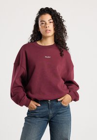 Vrouw met krullend haar, gekleed in een bordeauxrode sweatshirt met de tekst "New York" en blauwe jeans, staand met handen in haar zakken tegen een effen achtergrond.