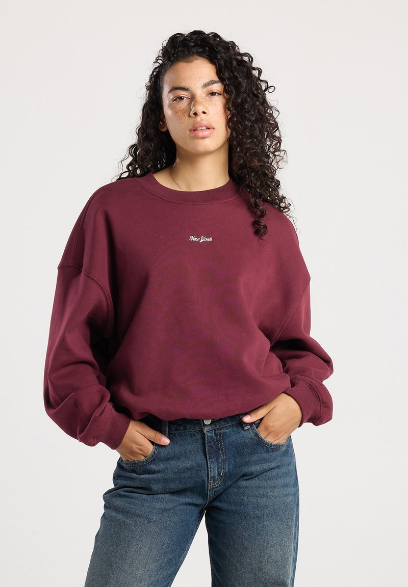 Vrouw met krullend haar, gekleed in een bordeauxrode sweatshirt met de tekst "New York" en blauwe jeans, staand met handen in haar zakken tegen een effen achtergrond.