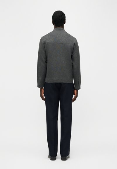 Άνδρας με την πλάτη στραμμένη, φοράει εφαρμοστό γκρι μακρυμάνικο μπουφάν και μαύρο παντελόνι ίσιας γραμμής, μπροστά σε απλό ανοιχτό φόντο.