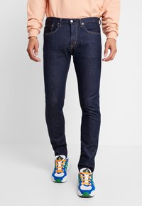 Man som bär åtstramade mörkblå jeans, en persikofärgad långärmad skjorta och blå, vita, gröna sneakers med orange snören står på ett vitt golv.