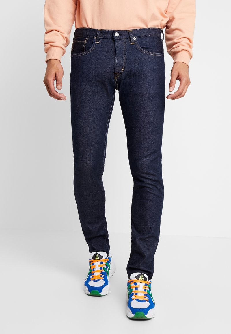 Man som bär åtstramade mörkblå jeans, en persikofärgad långärmad skjorta och blå, vita, gröna sneakers med orange snören står på ett vitt golv.