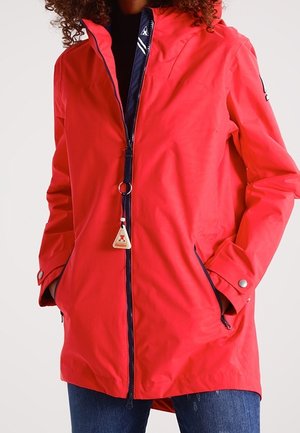 Kobieta nosząca jaskrawoczerwoną wodoodporną kurtkę na zamek błyskawiczny z przednimi kieszeniami, mankietami na zatrzaski i trójkątnym zawieszkiem na zamku.
