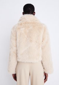 Veste en fausse fourrure beige clair avec un col haut, un design court et des manches larges, offrant une texture douce et des fibres longues semblables à des cheveux.
