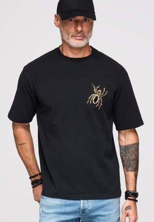 Mężczyzna w czarnej czapce, czarnym t-shircie z wzorem pająka w kolorze złotym i perłowym, jasnoniebieskich dżinsach oraz bransoletkach, z tatuażami na obu ramionach.