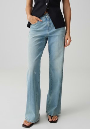 Donna con jeans azzurri a gamba larga leggermente consumati, gilet nero con bottoni e sandali neri con punta quadrata, in piedi con una mano in tasca.