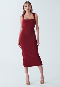 Bordeauxrotes figurbetontes Kleid mit quadratischem Ausschnitt, ärmellosem Design und dezentem seitlichem Schlitz; aus glattem Stoff gefertigt, kombiniert mit Riemchenabsätzen.