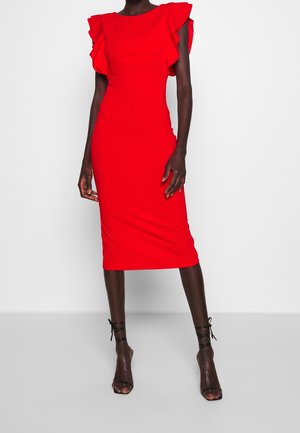 Mujer que lleva un vestido ceñido de color rojo brillante hasta la rodilla con mangas con volantes y sandalias negras de tacón alto con tiras, de pie sobre un fondo liso.