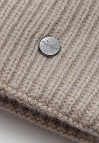 Béžová pletená čepice z vlny s žebrovaným vzorem, zdobená stříbrným kovovým knoflíkem vyrytým textem "PEARL WOOD". Měkká textura s viditelnými vlákny.