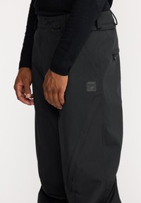 Pantalons imperméables noirs avec une finition texturée, comportant un patch logo, une taille réglable et des poches zippées sur les côtés. Coupe ajustée.