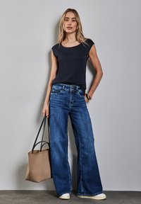 Námořnická modrá tričko s krátkým rukávem spárované se širokými modrými džíny a béžovou taškou. Oblečení se vyznačuje ležérním stylem s denimovou texturou a světlými odstíny.