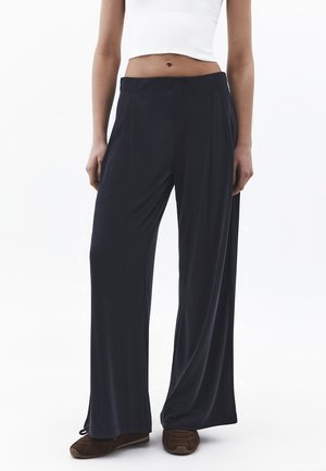 Persona che indossa pantaloni neri larghi e morbidi, top bianco senza maniche e corto, e scarpe casual marroni, in piedi contro uno sfondo bianco.