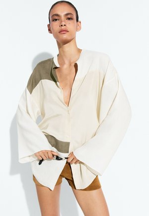 Femme aux cheveux tirés en arrière portant une chemise ample crème et olive et un short marron, ajustant une ceinture noire à la taille.