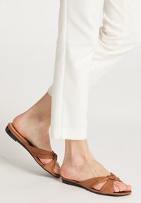 Sandali in pelle marrone a fascia con design annodato, suola piatta e punta aperta. Indossati con pantaloni bianchi a gamba dritta, che mostrano semplicità ed eleganza.