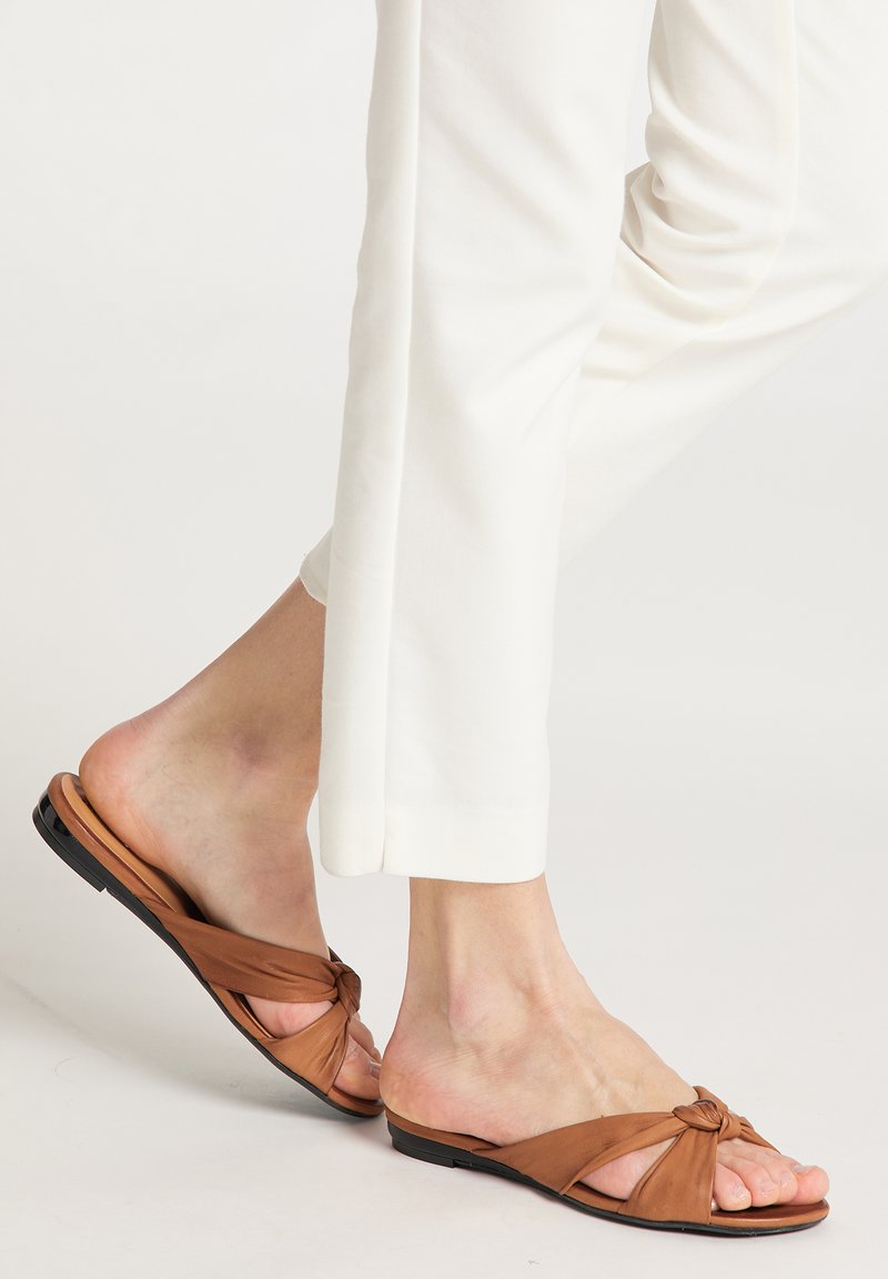 Sandali in pelle marrone a fascia con design annodato, suola piatta e punta aperta. Indossati con pantaloni bianchi a gamba dritta, che mostrano semplicità ed eleganza.