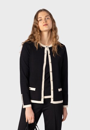 Jeune femme aux longs cheveux ondulés portant un cardigan noir avec une bordure blanche et un pantalon noir, tenant un sac à main noir sur un fond clair.