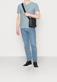 Černá kožená krosbodová taška s hladkým povrchem, vybavená zipem a logem, kterou nosí osoba v pruhovaném modrém tričku a světlých džínách.