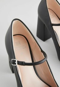 Scarpe nere con tacco alto, punta tonda, tacco a blocco e cinghietta alla caviglia. Esterno in pelle liscia e fodera beige morbida con dettaglio del logo.