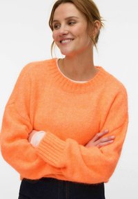 Femme aux cheveux châtain clair souriante, portant un pull orange vif en matière pelucheuse et un pantalon foncé, les bras croisés devant un fond blanc.
