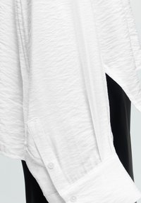 Poignet de chemise blanche texturée à manches longues avec détail de bouton, drapée sur un fond de pantalon noir.