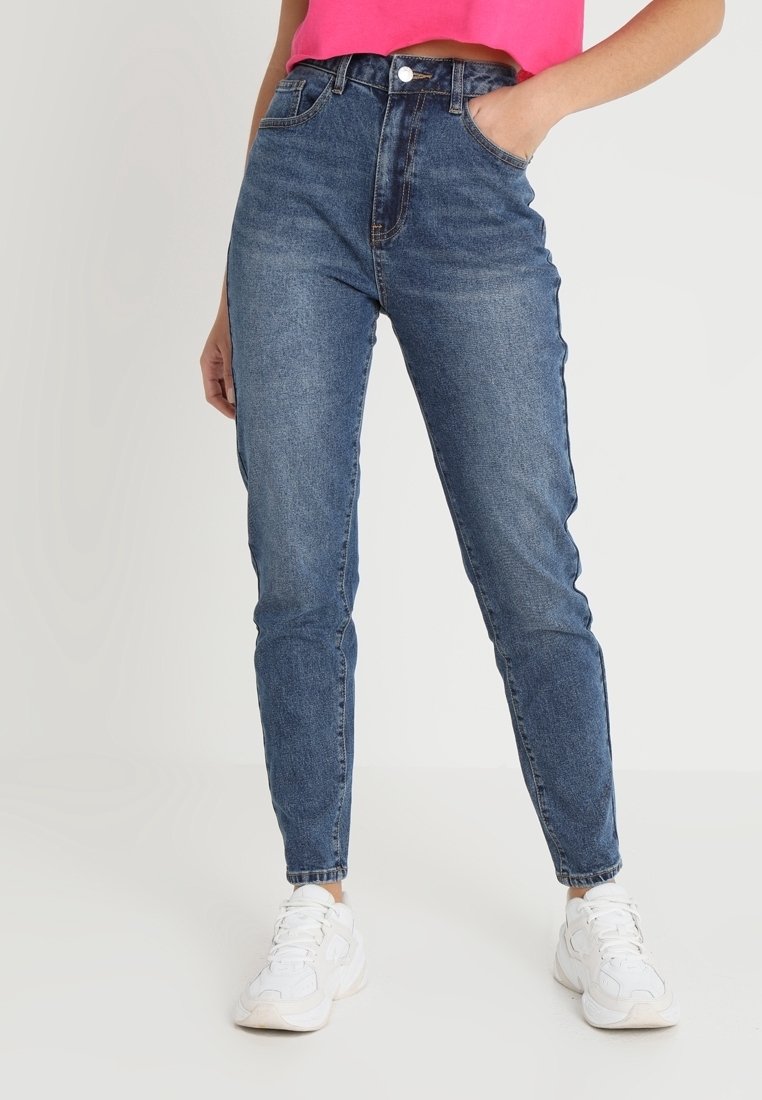 Femme portant un jean en denim bleu taille haute avec poches avant, associé à des baskets blanches et un crop top rose vif.
