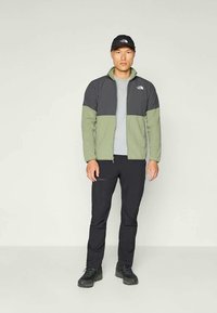 Lekka kurtka męska w kolorze oliwkowej zieleni i szarości, z zamkiem na przodzie, kontrastowymi panelami i dopasowanym krojem, zestawiona z czarnymi spodniami i butami.