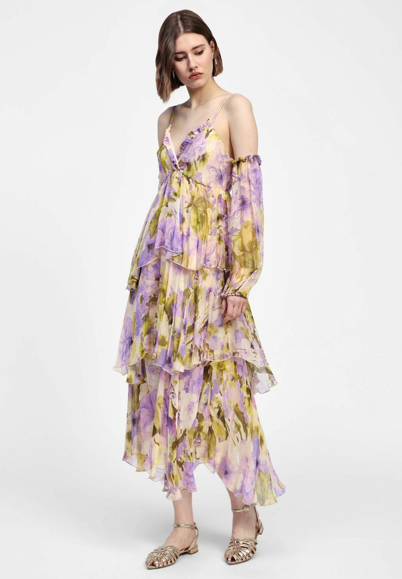 Abito maxi floreale in tessuto trasparente, con rouches sovrapposte, maniche scoperte sulle spalle e tonalità di viola e verde. Sandali argentati.